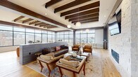 Located at 419 W Sixth St in Covington, Kentucky, this elevated amenity lounge is a thoughtfully designed communal space that blends contemporary style with warm, organic materials. The single-floor layout is anchored by dramatic exposed wood beam ceilings that contrast beautifully against crisp white soffits, creating a sense of architectural depth and character throughout.

Floor-to-ceiling wraparound windows form a defining feature of the space, flooding the interior with abundant natural light while offering sweeping panoramic views of the Covington skyline and surrounding cityscape, including visible bridges and neighboring rooftops. Light-toned hardwood flooring runs throughout, enhancing the bright, airy atmosphere.

The main lounge area is furnished with a rich leather sectional sofa, tufted accent chairs, and a curated mix of seating arrangements centered around a glass coffee table, all set atop a layered area rug. A stone-clad fireplace with a wall-mounted television anchors one end of the lounge, while globe pendant lights add a refined finishing touch.

A dedicated bar and kitchenette area features bold teal cabinetry with brass hardware, a mosaic tile backsplash, and a pass-through window overlooking the lounge. A separate high-top bar table with black counter stools provides additional casual seating. The overall ambiance is modern, inviting, and ideal for social gatherings or relaxed lounging.