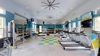 This modern fitness center is thoughtfully designed to support a wide range of workout routines in a bright and energizing environment. The space features high ceilings accented with recessed lighting and a large ceiling fan, ensuring excellent airflow and visibility throughout. A bold teal and white color scheme runs along the walls, lending the space a vibrant, motivating atmosphere.

The cardio zone is lined with a row of TRUE-brand treadmills positioned along one side of the room, each equipped with individual monitors for entertainment during workouts. On the opposite side, a well-stocked free weight area includes a full rack of dumbbells, a weight bench, and various functional training accessories such as medicine balls, resistance bands, and a stability ball. A dedicated stretching and functional training zone at the center of the room features a colorful checkered floor mat in yellow and blue, clearly delineating the space for floor-based exercises.

Large windows along the walls allow natural light to fill the room, while wall-mounted flat-screen televisions provide additional entertainment options. Full-length mirrors are strategically placed to assist with form and enhance the sense of space. The overall design balances functionality with an inviting aesthetic, making this fitness center a well-rounded facility for daily exercise.