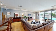 This single-floor community clubhouse is thoughtfully designed to offer a variety of functional gathering spaces within an open, flowing layout. The interior is unified by warm light hardwood flooring, soft blue-grey walls, and crisp white crown moulding throughout, creating a welcoming and cohesive ambiance.

The main lounge area is generously proportioned, furnished with large brown leather sectional sofas arranged around a rustic wood-and-metal coffee table. Track lighting overhead provides ample illumination, while large sliding glass doors along one wall flood the space with natural light and offer direct views of an outdoor pool and patio area.

Adjacent to the lounge, a formal dining zone features a long wooden dining table with high-back chairs, accented by decorative columns and arched doorways that lend a classic architectural character to the space.

A café-style kitchenette is equipped with light wood cabinetry, dark countertops, a coffee station, and a breakfast bar with woven rattan seating, serving as a convenient refreshment area.

A dedicated business center includes computer workstations and seating, positioned near bold framed artwork. Throughout the space, abundant greenery and curated décor contribute to an inviting, resort-like atmosphere.