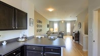 This three-floor home is designed with an open-concept layout on the main level, seamlessly connecting the kitchen, dining, and living areas into a cohesive and functional space. The kitchen features dark-finished cabinetry with sleek hardware and a generously sized island with a grey countertop, offering both prep space and casual seating. The island serves as a natural divider between the kitchen and the dining area, which is positioned to take advantage of the open sightlines throughout the space.

The living area is bathed in natural light from multiple windows dressed with grey patterned curtains, creating a bright and welcoming atmosphere. Light hardwood flooring runs throughout the main level, lending warmth and continuity to the space. The living room is comfortably appointed with ample seating and accented by decorative wall arrangements and soft ambient lighting from table lamps and a chandelier-style pendant light. A staircase is visible toward the rear, providing access to the additional floors above. Neutral wall tones throughout contribute to a clean, airy aesthetic well-suited for everyday living.