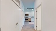This single-floor unit showcases a clean, contemporary design with a cohesive aesthetic carried throughout every room. Light hardwood-style flooring runs continuously across the space, complementing the crisp white walls and creating a bright, airy atmosphere.

The kitchen is a standout feature, equipped with full-height white flat-front cabinetry, white square tile backsplash, and a complete suite of stainless steel appliances including a range, microwave, dishwasher, and refrigerator. Track lighting overhead adds both function and a modern industrial touch, while the open layout allows easy flow into the adjacent living and dining areas.

The living area is bathed in natural light thanks to large floor-to-ceiling windows that offer views of the surrounding neighborhood and city skyline. The space is well-proportioned and versatile.

Two bedrooms provide comfortable retreats, each featuring large windows that invite natural light and outdoor views. The rooms are finished with the same light flooring and neutral tones found throughout the unit.

The bathroom is finished in an all-white palette with large vertically stacked wall tiles in the tub-shower surround, a wide vanity with an integrated sink, a large mirror, and chrome fixtures — delivering a sleek, spa-like feel. A central hallway connects all rooms efficiently, with additional storage accessible off the entry.