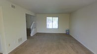 This two-story home presents a straightforward and functional layout designed to maximize livable space across both floors. The main level showcases a generously sized living area finished with wall-to-wall carpeting, providing a warm and comfortable foundation underfoot. Neutral-toned walls keep the space feeling bright and versatile, ready to accommodate a variety of interior styles. A centrally located window allows natural light to filter into the living area, offering a view of the outdoor surroundings, including glimpses of a fence and surrounding greenery. A staircase with white-painted wooden railings serves as a focal point near the entry, providing access to the upper level while contributing a clean, classic architectural detail to the main floor. The transition between the carpeted living area and the staircase landing is clearly defined, giving the layout a sense of organized flow. The upper floor continues the home's consistent design language. Overall, the space presents a practical and welcoming environment, with its neutral palette and carpeted interiors creating a move-in-ready feel suitable for a range of lifestyles.