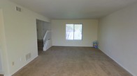 This two-story home presents a straightforward and functional layout designed to maximize livable space across both floors. The main level showcases a generously sized living area finished with wall-to-wall carpeting, providing a warm and comfortable foundation underfoot. Neutral-toned walls keep the space feeling bright and versatile, ready to accommodate a variety of interior styles. A centrally located window allows natural light to filter into the living area, offering a view of the outdoor surroundings, including glimpses of a fence and surrounding greenery. A staircase with white-painted wooden railings serves as a focal point near the entry, providing access to the upper level while contributing a clean, classic architectural detail to the main floor. The transition between the carpeted living area and the staircase landing is clearly defined, giving the layout a sense of organized flow. The upper floor continues the home's consistent design language. Overall, the space presents a practical and welcoming environment, with its neutral palette and carpeted interiors creating a move-in-ready feel suitable for a range of lifestyles.