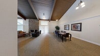 Located at 13222 E Iliff Ave in Aurora, CO, this single-story clubhouse offers a generously sized, open-concept gathering space designed for both casual socializing and organized meetings. The interior is immediately defined by dramatic vaulted ceilings with rich, dark wood planking and exposed beams, lending the space a warm, lodge-inspired character throughout.

At one end of the main hall, a towering floor-to-ceiling natural stone fireplace serves as a striking focal point, flanked by comfortable leather seating and a wood coffee table to create an inviting lounge area. Large windows with vertical blinds allow natural light to fill the space while maintaining privacy. Adjacent to the lounge, a formal conference table with upholstered chairs provides a dedicated area for meetings or private gatherings.

Transitioning toward the opposite end of the clubhouse, high-top bistro tables and barstool seating offer a casual dining setting, enhanced by pendant lighting suspended from the soaring ceiling. A pass-through bar counter connects this area to a separate kitchen equipped with modern appliances, including a stainless steel refrigerator, providing convenient service access for events.

Neutral carpet flooring, crown molding, and tasteful wall art contribute to a polished yet welcoming ambiance throughout, making this clubhouse a well-appointed venue for community use.