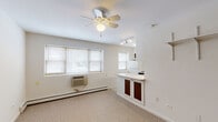 Located at 5746 N Kenmore Ave in Chicago's Edgewater neighborhood, this single-floor unit offers a clean, functional layout with a bright and neutral aesthetic throughout. The open living area features wall-to-wall carpet, a ceiling fan with light fixture, and multiple windows fitted with blinds that allow natural light to fill the space. A baseboard heating system and a wall-mounted AC unit provide year-round climate control. Adjustable wall-mounted shelving adds practical storage along one wall. The living area flows seamlessly into the kitchen via a peninsula counter with dark-accented cabinetry below, creating a natural divide between the two spaces. The kitchen is efficiently designed with white upper and lower cabinetry featuring dark wood-toned door panels, a gas range, refrigerator, stainless steel sink, and tile backsplash. Track lighting overhead illuminates the cooking and prep areas. The bathroom maintains a clean, classic look with white tile flooring, a vanity with under-sink storage, a large wall mirror with overhead lighting, and a shower-tub combination. The overall design is straightforward and well-maintained, offering a practical living environment in a convenient Chicago North Side location.