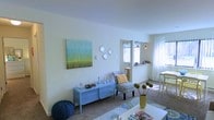 This single-floor residence offers a warm and inviting atmosphere defined by its light-filled interiors and thoughtfully curated décor. The living area serves as the heart of the home, featuring plush carpeting underfoot and soft, neutral walls that create a calm backdrop for colorful accents. A bold teal and green abstract canvas anchors one wall, complemented by decorative geometric mirror accents and a powder-blue console table styled with decorative objects. A patterned accent chair with a mustard yellow throw pillow adds a playful, eclectic touch, while a knit pouf and a low white coffee table complete the casual seating arrangement. Natural light streams in generously through sheer curtains, brightening the space throughout the day. An open pass-through or transitional area leads to a dining nook, where a bistro-style pendant light and a small table create an intimate setting for meals. Floral roman shades frame the windows, adding a charming finishing detail. The overall ambiance is relaxed and welcoming, blending soft tones with vibrant pops of color for a cheerful, lived-in feel that makes this space both functional and full of personality.