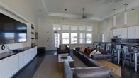 This single-floor space is designed as a comfortable and functional lounge area, characterized by its notably high ceilings that lend an open, airy atmosphere throughout. A ceiling fan is centrally mounted, enhancing both circulation and the room's visual height. Large, multi-pane windows span a generous portion of the wall, allowing abundant natural light to fill the interior while offering views to the outside.

The seating arrangement is anchored by a substantial dark leather sectional sofa and matching armchairs, all positioned around a rustic wooden coffee table atop a neutral area rug. Decorative throw pillows in warm tones add visual interest to the seating arrangement. Dark hardwood flooring runs throughout, providing a sleek, grounding contrast to the bright white walls and trim.

A built-in white shelving unit lines one wall, offering both display and storage space, and is complemented by framed artwork. Recessed lighting and track lighting supplements the natural light, ensuring the space remains well-lit at all times. Overall, the space presents a welcoming, well-appointed environment suited for gathering and relaxation.
