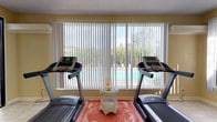 The fitness center at 2414 Stone Rd is a well-appointed, single-story exercise facility designed to support a comprehensive workout routine. The space is divided into two distinct zones, each thoughtfully equipped and finished to create a functional and comfortable environment.

The cardio area features light-toned tile flooring and warm yellow walls, creating a bright and energizing atmosphere. Two treadmills are positioned facing large windows fitted with vertical blinds, offering direct views of an outdoor swimming pool and lush surrounding trees. Natural light floods the room, enhancing the openness of the space.

A second, carpeted zone houses a variety of strength training equipment, including a multi-station cable and weight machine, a rowing bench, an elliptical trainer, and free weights. This area shares the same warm yellow wall tones and benefits from its own set of large windows that invite additional natural light. Vertical blinds provide adjustable light control throughout.

The two zones are connected by a glass partition with sliding doors, allowing the spaces to function as one open fitness area or to be divided as needed. The overall design balances practicality with comfort, offering a well-lit, ventilated, and versatile space suitable for both cardio and strength training activities.
