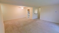 This single-floor space features a generously sized open living area finished with wall-to-wall neutral-toned carpeting, creating a clean and understated aesthetic throughout. The living area is outfitted with a ceiling fan, providing both light and air circulation. The layout flows seamlessly toward the kitchen, which is accessible through a recessed opening. The kitchen is equipped with white appliances, including a gas range and refrigerator, complemented by warm wooden cabinetry above. A pass-through counter or bar area connects the kitchen to the adjacent space, enhancing the open feel and allowing for easy interaction between the cooking and living areas. The walls throughout are painted in a soft, light neutral tone, contributing to a bright and airy atmosphere. The overall layout is practical and straightforward, with clear sightlines across the main living space that make it feel open and easy to navigate. The single-floor design ensures convenient accessibility throughout the unit, making it a functional and livable space.