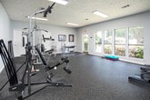 This single-floor fitness center offers a comprehensive workout environment designed to accommodate a variety of exercise routines. The space is divided into distinct functional zones, beginning with a strength training area that houses a multi-station cable machine, allowing users to perform a wide range of resistance exercises. Adjacent to this, a dedicated cardio section features elliptical machines and treadmills, all arranged on durable, speckled rubber flooring that provides both safety and comfort underfoot. A separate stretching and light exercise room offers a more open layout, equipped with an aerobic step platform and free weights, making it well-suited for flexibility training and low-impact workouts. Large windows line the walls of this area, flooding the room with natural light and providing pleasant views of the surrounding landscaping. A full-length mirror in the main workout area visually expands the space and allows users to monitor their form. Framed artwork adorns the walls, adding a polished touch to the functional environment. Textured ceilings with recessed fluorescent lighting ensure consistent, bright illumination throughout. French doors connect the fitness center to an adjacent common area, enhancing the sense of openness. The overall ambiance is clean, bright, and purpose-built for an active lifestyle.