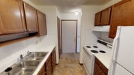 This single-floor residential unit offers a practical and straightforward layout designed for comfortable everyday living. The galley-style kitchen is efficiently organized with warm wood cabinetry on both sides, white laminate countertops, a double stainless steel sink, and a white coil-top electric range with an overhead exhaust hood. Tile flooring runs through the kitchen, which leads directly to the unit's entry door, creating a functional transition space.

The living area is carpeted and bathed in natural light through a large sliding glass door dressed with vertical blinds, which provides access to an outdoor space. A wall-mounted air conditioning unit and a baseboard heater ensure year-round climate comfort. The open, neutral-toned walls give the room a clean and versatile feel.

The bedroom is similarly carpeted and features a window with horizontal blinds that allows natural light to filter in. A textured ceiling and baseboard heating are consistent throughout the sleeping areas, contributing to a cohesive interior finish.

The bathroom includes a full-size soaking tub and shower combination with a curtain rod, a toilet, and a wall-mounted medicine cabinet with a mirror. Tile flooring and cream-toned walls complete the space with a clean, functional aesthetic throughout.