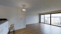 This single-floor space offers a clean, functional layout with a bright and open living area at its core. The flooring throughout is finished in light-toned wood, lending a warm and welcoming feel to the interior. A standout decorative element in the living area is the gold-toned chain-hung pendant light, which adds a classic touch to the otherwise simple, neutral aesthetic.

The walls are finished in a textured white, providing a neutral backdrop that maximizes the sense of light and openness throughout the space. Natural light flows generously into the main living area through a large sliding glass door, which opens onto a private balcony. The balcony features a railing and overlooks a surrounding outdoor environment, offering pleasant exterior views and a connection to the outdoors.

The open layout of the main living area provides flexible use of space, suitable for a variety of furniture arrangements. The combination of natural light, light-toned flooring, and neutral wall finishes creates a bright, airy ambiance throughout. Overall, the space presents a straightforward and adaptable interior with practical design elements and a comfortable, light-filled atmosphere.