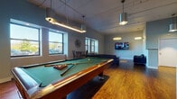 Located at 1317 East St in New Britain, Connecticut, this single-floor community lounge and recreation room is designed to offer residents a well-rounded social and entertainment experience. The space features warm hardwood-style flooring throughout, complemented by cool blue-gray walls that give the room a modern, relaxed aesthetic.

At the center of the space sits a full-size pool table with a green felt surface, positioned beneath a cluster of sleek, cylindrical pendant lights that provide focused illumination. A wall-mounted mirror nearby enhances the sense of depth and openness in the room.

The lounge area is furnished with a large black leather sectional sofa and a pair of tufted leather armchairs arranged around a flat-screen television mounted on the wall, creating an inviting space for watching shows or simply relaxing. Natural light streams in through large, deep-set windows, offering views of the surrounding outdoor greenery.

A bistro-style table with metal chairs provides a casual dining or conversation nook near the windows. Additional amenities include a beverage vending machine for added convenience. The space is finished with recessed and track lighting throughout, ensuring the entire area is well-lit. Overall, the room is thoughtfully designed to serve as a versatile communal hub for recreation and relaxation.