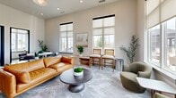 Located at 1801 Walt Whitman Dr, this thoughtfully designed single-floor space combines a welcoming lounge with two dedicated private offices. The living room serves as the heart of the space, boasting soaring ceilings and floor-to-ceiling windows fitted with roller shades that bathe the interior in generous natural light while offering open views of the surrounding outdoor landscape. The room is furnished with a rich cognac leather sofa, olive-green swivel lounge chairs, a sculptural dark wood oval coffee table, and cane-back counter-height chairs arranged along a low credenza — creating a warm, contemporary atmosphere. Patterned low-pile carpeting in soft gray tones flows throughout, tying the aesthetic together cohesively. Lush potted olive trees and moss-topped decorative bowls introduce organic accents that complement the neutral, earthy palette. The two private offices are each equipped with a clean-lined white desk, a monitor workstation, and task lighting, with artwork adorning the walls and windows providing views to the outdoors. The overall ambiance is polished yet inviting, balancing productivity and comfort in a refined, modern setting.