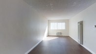 This single-floor space offers a clean, minimalist layout designed for comfortable everyday living. The main living area is a standout feature, showcasing rich, dark hardwood-style flooring that contrasts beautifully against the crisp, neutral-toned walls. A large window dressed with vertical blinds allows an abundance of natural light to flood the room, creating a bright and airy atmosphere throughout the day. The textured ceiling adds a subtle architectural detail overhead, while white baseboards provide a polished finish along the perimeter of the room. A wall-mounted air conditioning unit is conveniently positioned beneath the window, ensuring year-round climate comfort. The open layout of the living area provides a versatile canvas, easily adaptable to a variety of furniture arrangements and personal styles. The overall ambiance of the space is calm and understated, with a neutral color palette that lends itself well to a wide range of interior design preferences. The combination of natural light, durable flooring, and an open floor plan makes this a practical and inviting space suitable for a range of uses.