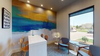 Located along Grand Cordera Parkway in Colorado Springs, Colorado, this single-floor business center offers a thoughtfully designed workspace that balances functionality with contemporary style. The space is immediately distinguished by a striking large-scale abstract mural rendered in bold sweeps of gold, teal, and deep blue, serving as a dramatic focal point that lends an artistic energy to the room.

The business center is organized into distinct functional zones. A dedicated computer workstation area features multiple iMac desktop computers arranged along a built-in counter with a marble-style surface and dark cabinetry, complemented by ergonomic seating and a multifunction printer. Recessed lighting throughout ensures consistent illumination across all work surfaces.

A collaborative seating area includes a sleek white high-top table with bentwood stools, ideal for informal meetings or individual work. Adjacent to this is a lounge-style corner outfitted with mid-century-influenced upholstered chairs and a round side table, positioned beside a tall floor-to-ceiling window that floods the area with natural light and provides views of the surrounding landscape. Patterned carpet in blue-gray tones ties the space together, creating a cohesive, professional atmosphere suited for productivity.