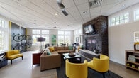 The District Community Center in University City, Missouri offers a thoughtfully designed single-floor layout that balances functionality with comfort. Upon entry, a warm reception area greets visitors with a substantial wooden desk, patterned carpet tiles in earthy tones, and an adjacent private office behind a glass-panel door.

The central lounge serves as the heart of the space, anchored by a dramatic floor-to-ceiling stacked stone fireplace with a mounted flat-screen television above. Generous seating, including a large sectional sofa and bold yellow accent chairs, invites residents to gather and relax. Ceiling fans and abundant natural light from large, multi-pane windows lend the room an airy, open atmosphere with pleasant outdoor views.

Adjacent to the lounge, a bright dining area features bistro-style round tables with black chairs and expansive windows that flood the space with daylight. A workstation with a desktop computer provides a practical resource for residents.

Two kitchenette areas are equipped with rich wood cabinetry, granite countertops, stainless steel appliances, and tile backsplashes, offering convenient amenities for community use. A transitional area with polished concrete flooring and clerestory windows connects the zones seamlessly, completing this well-appointed community gathering space.