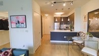 This single-floor space showcases a thoughtfully designed open-concept layout that seamlessly connects the living, dining, and kitchen areas. The kitchen is a standout feature, outfitted with dark espresso cabinetry, a light-toned countertop island with a built-in sink, and stainless steel appliances including a full-sized refrigerator. A series of sleek pendant lights hang above the island, complementing the track lighting overhead that illuminates the space with a warm, modern glow. The light hardwood-style flooring runs consistently throughout, adding warmth and visual continuity to the interior. White walls and clean-lined doorways contribute to a crisp, airy ambiance, while subtle decorative touches — including a colorful wall piece in the living area — add personality to the space. The entryway is clearly defined and functional, providing a smooth transition into the main living areas. Overall, the space strikes a balance between contemporary design and practical living, making it both visually appealing and highly functional for everyday use.