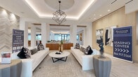 This single-floor space showcases a seamlessly connected series of modern, well-appointed areas designed for both comfort and sophistication. The main lounge is a standout feature, anchored by a striking geometric chandelier suspended beneath a coffered ceiling with warm cove lighting. Crisp white sofas, a round marble coffee table, and curated sculptural artwork establish an elevated, gallery-like atmosphere. Herringbone-patterned light wood flooring flows throughout, tying each area together cohesively.

A secondary lounge zone offers a large built-in entertainment wall clad in warm wood paneling, complemented by integrated display niches with accent lighting. Comfortable seating arrangements, including cognac leather chairs and upholstered benches, face the media wall, while floor-to-ceiling glass doors open directly to an outdoor area, flooding the space with natural light and exterior views.

The kitchen is meticulously designed with a generous island featuring a white stone countertop and built-in cooktop, warm wood cabinetry, and a dramatic textured tile accent wall housing the sink station. Stainless steel appliances and recessed lighting add a clean, professional quality to the space. An adjacent dining area accommodates a large communal table beneath a trio of conical pendant lights, with expansive glazed walls framing lush outdoor greenery, creating a bright and inviting setting for gathering.