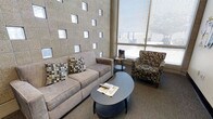 This single-floor space presents a thoughtfully designed lounge area that balances architectural character with functional comfort. The most striking feature is a tall, textured accent wall adorned with a grid of square geometric cutouts, creating a visually dynamic backdrop that adds depth and a modern aesthetic to the room. Opposite the accent wall, expansive floor-to-ceiling windows fitted with horizontal blinds flood the space with natural light and offer views of the surrounding exterior, enhancing the open and airy atmosphere.

The seating arrangement is both inviting and practical, centered around a neutral-toned sofa accented with patterned throw pillows in earthy tones. A pair of upholstered accent chairs with a matching mosaic-style pattern complement the sofa and contribute to the cohesive design palette. A uniquely shaped, kidney-style coffee table in a muted blue-grey finish serves as the focal point of the seating arrangement, providing a functional surface for reading materials. A small side table rounds out the furniture grouping. The carpeted flooring in a neutral grey tone ties the elements together, making this space well-suited as a waiting area, reception lounge, or collaborative gathering space.