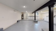 This single-floor utility space is designed for practical, everyday use and is thoughtfully laid out to accommodate shared laundry needs. The room is finished with durable, easy-to-clean grey epoxy flooring that extends throughout the entire area, providing a resilient surface well-suited for a high-traffic environment.

A row of commercial-grade top-load washing machines and dryers lines the far wall, offering ample laundry capacity. A small window near the machines allows for a degree of natural light to enter the space. Recessed ceiling lighting ensures the area is well-illuminated throughout.

The room also houses visible utility infrastructure, including a gas-powered water heater and a network of exposed pipes along one wall, underscoring the functional nature of the space. A bold black structural beam runs overhead and connects to a central support column, adding an industrial character to the otherwise clean, minimal aesthetic.

The open floor plan provides generous circulation space, making it easy to move around and manage laundry tasks. Concrete block benches are positioned within the space, offering convenient surfaces for sorting or folding. The overall ambiance is utilitarian and well-maintained, reflecting a space built for efficiency and durability.