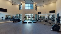 This well-appointed fitness center is defined by its striking architectural design, most notably the soaring double-height ceilings that create an open, airy atmosphere throughout the main exercise floor. Floor-to-ceiling windows with sleek black-framed grid patterning line the exterior walls, flooding the interior with natural light and offering views of the surrounding outdoor landscape.

The main cardio area is equipped with multiple treadmills, elliptical trainers, stationary bikes, and a rowing machine, all arranged to face the large windows. Ceiling-mounted fans provide additional ventilation and comfort. Wall-mounted flat-screen televisions are positioned at strategic heights for visibility during workouts. Full-length mirrors along one wall enhance the sense of space and allow users to monitor their form.

A dedicated strength and functional training zone features adjustable weight benches, kettlebells, medicine balls, stability balls, and a dumbbell rack. A separate, more enclosed area is outfitted with suspended TRX suspension training straps anchored to the ceiling, offering a versatile space for bodyweight and resistance-based workouts.

Throughout the facility, durable rubber flooring provides safety and comfort. Recessed and pendant lighting supplements the natural light, ensuring consistent illumination. The overall design is clean, modern, and functional.