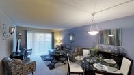 This single-floor residence features an open-concept layout that seamlessly integrates the dining and living areas into one cohesive space. The living room is anchored by a plush carpet and styled with a dark leather sofa, a patterned area rug, and a decorative sunburst mirror mounted on a cool blue accent wall. A large sliding glass door with vertical blinds floods the space with natural light while offering outdoor views and access.

Adjacent to the living area, the dining space is defined by a sleek glass-top table surrounded by white upholstered chairs, illuminated by a modern pendant light fixture that hangs from the textured ceiling. A sideboard along the wall provides additional storage and display space, accented by framed artwork and decorative accessories.

The textured ceiling adds a classic architectural detail throughout, while the combination of warm yellow tones on select walls and the cool blue accent wall creates a visually dynamic yet cohesive atmosphere. The open layout enhances the sense of spaciousness, making the area well-suited for both everyday living and entertaining.