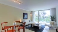 This stylish single-floor space showcases a seamlessly integrated living and dining area filled with natural light streaming through expansive floor-to-ceiling windows. The windows open to a balcony overlooking lush greenery and neighboring architecture, creating a vibrant indoor-outdoor connection. Crown molding adds a refined architectural touch to the smooth white ceilings, which are complemented by a sleek track lighting system overhead. The living area is anchored by a crisp white sofa and a modern rectangular coffee table atop a dark, patterned area rug, lending contrast and depth to the light-toned space. A bold abstract painting mounted on the warm beige wall serves as a striking focal point, while a sculptural mid-century-inspired arc floor lamp adds a designer flair. The dining area features a compact round glass-top table paired with distinctive burnt-orange chairs, introducing a vibrant pop of color against the neutral palette. White tile flooring runs throughout, enhancing the airy and clean aesthetic of the space. Overall, the residence strikes a balanced blend of contemporary design and comfort, with thoughtful furnishings and an abundance of natural light creating a welcoming and visually engaging environment.