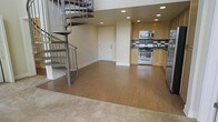 This two-story space is defined by an open-concept main level that integrates the living and kitchen areas into a cohesive, functional layout. The kitchen is outfitted with light maple cabinetry, stainless steel appliances including a gas range and built-in microwave, and a dark geometric tile backsplash that adds a contemporary accent. Recessed lighting throughout the ceiling provides consistent, even illumination across the space.

Smooth wood-toned flooring extends across the main living area, transitioning to carpet at the perimeter, creating a subtle distinction between zones. Neutral-toned walls keep the interior feeling clean and versatile.

A standout architectural feature is the metal spiral staircase positioned near the entrance, which serves as both a functional element and a visual focal point, connecting the main level to the upper floor. The staircase's industrial design contrasts tastefully with the warm cabinetry and flooring tones. The overall ambiance is modern and understated, with a practical layout that makes efficient use of the available space across both levels.