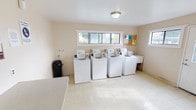 The Harris Place Laundry Facility is a dedicated, single-floor shared laundry room designed to serve the needs of its residents efficiently and conveniently. The space is thoughtfully laid out to accommodate multiple users at once, featuring a row of four top-loading washing machines along one wall and two stacked washer-dryer units along another, providing ample capacity for laundry needs. The room is finished with durable vinyl tile flooring in a neutral speckled pattern, which is both practical and easy to maintain. Warm, light-toned walls and flush-mount ceiling lighting keep the space bright and functional. A built-in folding counter runs along one wall, offering a dedicated surface for sorting and folding laundry. A pair of metal chairs provides seating while waiting. Natural light filters into the room through a set of horizontal windows positioned high on the wall, offering views of surrounding greenery. A bulletin board and posted signage, including the facility's usage rules and guidelines, keep residents informed. The room also includes a waste bin for convenience. The overall layout is clean, organized, and utilitarian, making it a practical shared amenity for the community.
