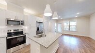 This thoughtfully designed single-floor residence offers a cohesive open-concept layout that seamlessly connects the kitchen and living area. The kitchen serves as the heart of the home, showcasing granite countertops, crisp white shaker cabinetry with subway tile backsplash, and a full suite of stainless steel appliances including a French door refrigerator, electric range, and built-in microwave. A central island with an undermount sink and stylish pendant lighting provides additional prep space and a natural gathering point. The adjoining living area is bright and airy, benefiting from large windows and a glass door that invite natural light and offer outdoor views. Warm hardwood-style flooring flows continuously throughout the entire unit, anchored by clean white baseboards and crown molding details. The spacious bedroom features a ceiling fan, a generously sized window with outdoor views, and access to a dedicated closet. The bathroom is finished with granite-top white vanity cabinetry, brushed nickel hardware, and recessed lighting. A hallway connects the private areas of the unit, maintaining a logical and functional flow. The overall palette of neutral tones, recessed lighting, and consistent finishes gives the space a clean, modern ambiance.