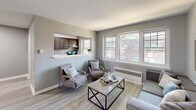 Located at 1120 N 66th St in Philadelphia's 19151 zip code, this 2-bedroom, 1-bathroom unit offers a thoughtfully designed single-floor layout with clean, contemporary finishes. The open living room is bathed in natural light from a generous bank of windows overlooking the neighborhood, and features light wood-look flooring and a neutral gray color palette that creates a calm, cohesive atmosphere. A pass-through opening connects the living area to the kitchen, which is equipped with dark cabinetry, a stainless steel refrigerator, and light countertops. The kitchen flows into a separate dining area, providing a functional and social layout. A hallway with an elegant arched doorway connects the main living spaces to the two bedrooms. The spacious primary bedroom accommodates a full-size bed comfortably, benefits from natural light through dual windows, and includes closet space. The second bedroom is a well-proportioned, versatile room with the same warm flooring. The full bathroom features a tub-shower combination with striking floor-to-ceiling marble-style tile, lending an elevated, clean aesthetic. Consistent light hardwood flooring, crisp white trim, and soft gray walls unify the entire unit with a modern, move-in-ready appeal.