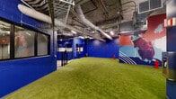 Located at 30 Peabody Street in Nashville, TN, this energetic pet amenity facility is purpose-built for dogs and their owners. The space is spread across a single floor and is divided into two primary functional areas connected by a secure entry with a double-gated airlock system to safely manage pet access.

The main play area features lush artificial turf covering a generously sized open floor, enclosed by bold cobalt-blue painted concrete block walls. Exposed ductwork and industrial-style ceilings lend the space an urban warehouse character, while vibrant large-scale murals depicting dogs in dynamic, colorful compositions bring energy and personality to every corner. Decorative red fire hydrants are placed throughout, adding a playful, thematic touch. Stainless steel waste stations are conveniently positioned for easy cleanup.

Adjacent to the play area, the dedicated dog wash room is thoughtfully designed with patterned blue-and-white geometric floor tiles, full-height white subway tile wainscoting, and multiple elevated wash bays equipped with handheld showerheads and hose reels. A wall-mounted dryer and utility sink complete the grooming setup. The wash room's bright, clean aesthetic contrasts beautifully with the play area's bold industrial style, together creating a comprehensive and visually engaging environment for pet care.
