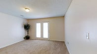 This single-floor space presents a straightforward and functional layout designed for comfortable everyday living. The main living area is finished with wall-to-wall beige carpeting that lends a warm, understated feel underfoot, complemented by smooth, neutral-toned walls that create a clean and bright atmosphere throughout.

A standout feature of the room is the set of white-framed French doors, which serve as both an architectural focal point and a source of natural light. The sidelights flanking the doors allow daylight to filter into the interior, enhancing the open and airy quality of the space. The doors provide direct access to an exterior area, adding a sense of connectivity between indoors and outdoors.

The ceiling features a classic textured finish and is equipped with a flush-mounted light fixture, providing consistent overhead illumination. White baseboards run along the perimeter of the room, offering a clean contrast against the carpeted floor. The room's simple, open layout makes it highly adaptable for a variety of furniture arrangements and uses. A decorative potted plant in the corner adds a natural, organic touch to the otherwise neutral interior.