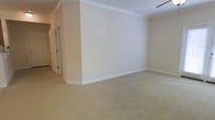 This single-floor space presents a generously sized main room finished with soft, neutral-toned walls that create a bright and calming atmosphere. The room is adorned with classic crown molding along the ceiling, adding a touch of traditional elegance to the otherwise understated design. Plush, light-colored wall-to-wall carpeting covers the floor, contributing to a warm and comfortable feel underfoot. A ceiling fan is partially visible, suggesting practical climate control within the space. Plantation shutters on the windows provide both privacy and light management, allowing natural light to filter in softly. Multiple electrical outlets are positioned along the walls, offering functional versatility for various furniture arrangements. White baseboards frame the perimeter of the room cleanly. A hallway visible through a paneled door connects to additional areas of the space, hinting at further rooms beyond. Overall, the space conveys a tidy, move-in-ready character with classic architectural detailing and a neutral palette suitable for a wide range of uses.