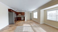 This single-floor, one-bedroom, one-bathroom unit offers an open-concept layout that seamlessly connects the kitchen and living areas. The kitchen is a standout feature, showcasing rich dark-wood cabinetry, a tasteful tile backsplash, stainless steel appliances including a refrigerator and dishwasher, and a small peninsula with built-in shelving that provides additional storage and prep space. Recessed lighting throughout the kitchen ensures ample illumination, complemented by crown molding that adds a refined architectural detail to the space.

The living area extends generously from the kitchen, finished with light-toned hardwood-style flooring that transitions smoothly throughout the open space. A large window along the far wall allows natural light to pour in, casting bright patterns across the floor and creating a warm, inviting ambiance. Neutral, cream-toned walls enhance the airy feel of the room. A doorway visible from the main area leads to the private bedroom, maintaining a practical and efficient flow throughout the unit.