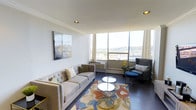 This well-appointed single-floor residence exudes a modern, sophisticated charm throughout its thoughtfully designed living space. The living room serves as the centerpiece of the home, featuring large floor-to-ceiling corner windows that flood the interior with abundant natural light and frame panoramic views of the surrounding cityscape and rolling hillside beyond. Crown molding along the ceiling adds a refined architectural detail, complementing the clean white walls and dark hardwood flooring that ground the room with a sense of warmth and contrast. The space is furnished with a tufted light grey sofa accented by decorative pillows, paired with a sleek grey wingback chair and a coordinating blue ottoman, creating an inviting and cohesive seating arrangement. A set of mirrored circular coffee tables rests atop a vibrant, multicolored area rug, introducing an artistic focal point to the room. Recessed lighting provides additional illumination, enhancing the airy atmosphere. A large abstract canvas on the wall adds a contemporary artistic touch, while a yellow floral accent near the window brings a subtle pop of color. The overall ambiance is bright, modern, and elegantly curated.