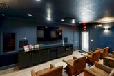 Located in Birmingham, Alabama, this intimate private movie theater delivers an upscale cinematic experience within a thoughtfully designed single-room space. The interior is defined by deep navy blue walls paired with a matte black textured ceiling, creating an immersive, light-controlled atmosphere ideal for screening. Recessed ceiling lights and warm wall sconces provide layered ambient lighting, enhancing the room's sophisticated ambiance without detracting from the viewing experience.

Seating consists of rows of generously proportioned brown leather club chairs with studded detailing, arranged on a gently tiered floor to ensure clear sightlines from every position. Small round side tables are conveniently placed throughout the seating area for guests' use. Framed cinema-themed blueprint artwork adorns the back and side walls, adding a stylish, thematic touch to the décor.

The focal point of the room is a large flat-screen display mounted above a sleek black cabinet unit, which houses a microwave and additional storage. A dedicated snack and refreshment station sits alongside, complete with curated snack options. Light wood-tone flooring contrasts cleanly with the dark walls and ceiling, grounding the space with warmth. This well-appointed theater offers a premium, private viewing environment suited for an engaging entertainment experience.