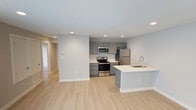 This single-floor residence offers a clean, contemporary design with an open-concept layout that maximizes the sense of space and flow. The kitchen is a standout feature, showcasing gray shaker-style cabinetry paired with a neutral subway tile backsplash and a full suite of stainless steel appliances, including a range, microwave, and refrigerator. A peninsula island with a white quartz-style countertop and an integrated sink provides additional workspace and casual seating, seamlessly connecting the kitchen to the main living area. Light hardwood-style flooring runs throughout the space, contributing to a bright and airy atmosphere. Recessed lighting is installed throughout, ensuring consistent, well-distributed illumination across all areas. The walls are finished in a soft, neutral gray tone that complements the overall modern palette. Wide doorways and an open floor plan create a sense of continuity between rooms. A hallway visible from the main living area leads to additional rooms, suggesting a thoughtfully designed and functional layout. The overall ambiance is fresh, modern, and move-in ready, appealing to those who appreciate clean lines and contemporary finishes.