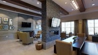 This single-floor space showcases a thoughtfully designed interior that blends contemporary style with warm, inviting elements. The centerpiece of the room is a dramatic floor-to-ceiling stacked stone fireplace column, which serves as a natural divider between functional areas while anchoring the space with a bold architectural statement. A linear gas fireplace insert is built into the base, adding both ambiance and practicality. Mounted flat-screen televisions on either side of the column ensure visibility from multiple vantage points throughout the room.

Exposed wooden ceiling beams run overhead, complementing the natural stone textures and adding a rustic yet refined character to the space. Recessed lighting and track fixtures provide layered illumination throughout. The lounge area features comfortable seating arranged around a patterned area rug, while a separate dining area with a dark wood table and upholstered chairs offers a more formal setting. Large windows allow natural light to filter in, creating a bright and open atmosphere. The combination of stone, wood, and neutral tones gives the overall space a cohesive, sophisticated ambiance suited for both relaxation and gathering.