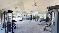 Located at 1520 N Beckley Ave in Dallas, Texas, this well-appointed fitness center offers a bright, open environment designed to support a comprehensive workout experience. The space is defined by soaring vaulted ceilings fitted with recessed lighting and dual ceiling fans, creating excellent airflow and an airy atmosphere throughout. Large floor-to-ceiling windows and glass panels line the perimeter, flooding the interior with abundant natural light and offering unobstructed views of the surrounding landscaped outdoor areas, including a pool and mature trees. The flooring features durable, multi-toned carpet tiles suited for high-traffic fitness use. A striking feature wall showcases a bold black-and-white geometric arch mural, adding a modern artistic touch that sets the space apart. The room is equipped with a diverse selection of cardio and strength-training equipment, including treadmills, ellipticals, stationary bikes, cable machines, weight benches, and a dumbbell rack with mirror backing. The layout is thoughtfully organized to allow ample room between stations, supporting safe and efficient movement. Crown molding along the upper walls adds a refined architectural detail, balancing the gym's functional design with a polished aesthetic. This fitness center provides a well-lit, visually engaging, and fully equipped environment for residents.