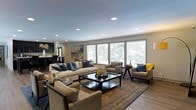 This thoughtfully designed single-floor residence showcases an open-concept layout that seamlessly connects the living and kitchen areas, creating a welcoming and functional flow. The expansive living room is the centerpiece of the home, anchored by a large area rug and a dark-finished coffee table that grounds the neutral, sophisticated furnishings. A generous arrangement of cream-toned sofas and accent chairs — accented with navy and yellow throw pillows — creates an inviting gathering space with a balanced, contemporary aesthetic. Recessed lighting in the smooth ceiling provides even illumination throughout, complementing the ample natural light that streams in through a row of wide windows dressed with horizontal blinds. The windows frame lush outdoor greenery, bringing a sense of the outdoors inside. In the background, a dark-cabinetry kitchen with pendant lighting adds contrast and depth to the open floor plan, hinting at a well-equipped culinary space. Hardwood-style flooring runs throughout, tying the spaces together cohesively. The overall ambiance is modern yet warm, offering a comfortable and stylish environment well-suited for both everyday living and entertaining.