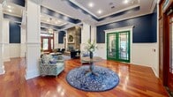 Located at 1 Legacy Oaks Pl in Asheville, NC, this single-floor living room is a showcase of refined craftsmanship and thoughtful design. Rich cherry hardwood floors stretch across the generously sized space, reflecting the abundant natural light that pours in through stately wood-framed French doors. The walls are dressed in a bold navy blue above crisp white wainscoting panels, creating a striking yet sophisticated color palette throughout.

The coffered ceiling is a standout architectural feature, with white trim accenting the navy insets and recessed lighting, while an elegant globe chandelier serves as a central focal point. White fluted columns define distinct areas within the open layout, lending a classical structural elegance to the room.

The floor-to-ceiling stacked stone fireplace commands attention as the room's anchor, complete with a rustic wood mantel and a raised stone hearth — ideal for Asheville's cooler mountain evenings. A bold green-framed glass door adds a vivid accent and connects to an adjacent area, while a secondary set of French doors opens to views of lush greenery outside.

The space is thoughtfully furnished with tufted velvet seating and layered blue area rugs, enhancing the cohesive, upscale aesthetic. Overall, this living room exudes a warm, welcoming ambiance with a distinctly classic Southern charm.