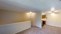 This single-floor residential space offers a straightforward and functional layout designed for comfortable everyday living. The main living area features wall-to-wall carpet in a neutral tone, contributing to a warm and inviting atmosphere. Textured popcorn ceilings are consistent throughout, and a central ceiling light fixture provides ambient illumination to the space.

A low half-wall or railing feature defines the boundary of the living area, adding subtle architectural interest while maintaining an open sightline into the adjacent kitchen. The transition between spaces is seamless, making the overall layout feel connected and cohesive.

The kitchen is visible from the living area and is equipped with dark wood cabinetry, both upper and lower, offering ample storage. A light-toned countertop contrasts with the cabinetry, and a built-in appliance is integrated beneath the counter. The kitchen's flooring transitions from the carpeted living space to a harder surface, providing a practical distinction between the two areas.

The walls throughout are painted in soft, warm beige tones, creating a consistent and neutral palette that suits a variety of furnishing styles. The overall ambiance is modest and utilitarian, making the space practical and easy to personalize.