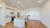 This single-story, 1-bedroom, 1-bathroom residence offers a thoughtfully designed open-concept layout. The kitchen serves as the centerpiece of the living area, showcasing a large island with granite countertops and a built-in sink, white shaker-style cabinetry, stainless steel appliances including a refrigerator and range, and recessed lighting overhead. Warm hardwood-style flooring extends throughout the main living and kitchen areas, adding a cohesive and inviting feel. A sliding glass door provides access to an outdoor balcony, allowing natural light to fill the space.

The bedroom is a comfortable, carpeted retreat featuring dual windows with outdoor views, a ceiling fan, and a clean, neutral palette. A second carpeted room offers additional flexibility for storage or other uses.

The bathroom is spacious and well-equipped, featuring granite-topped dual vanities with ample cabinetry, a large mirror, and a full tub-shower combination with white tile surround and accessibility grab bars. Hardwood-style flooring continues into the bathroom, tying the unit's design together cohesively.

Throughout the unit, crisp white walls, recessed lighting, and modern brushed-nickel hardware contribute to a clean, contemporary aesthetic that is both functional and visually appealing.