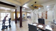 This two-story office space combines classic architectural elegance with practical, professional functionality. The main level immediately impresses with its striking dark wood fluted columns that rise from tiled flooring, adding a formal, distinguished character to the workspace. A decorative palm-leaf ceiling fan serves as a focal point overhead, complementing the recessed lighting and tray ceiling detail that adds depth and dimension to the space.

The layout is thoughtfully open, with clear sightlines connecting multiple work areas. French-style glass-paned doors partition private office spaces while maintaining a sense of openness and allowing natural light to flow throughout. The warm, neutral color palette on the walls and tile flooring creates a welcoming yet professional atmosphere.

Workstations are equipped with ergonomic seating and ample desk surfaces, accommodating collaborative or individual work arrangements. Additional areas visible beyond the columns suggest dedicated spaces for meetings or focused work, each separated by the elegant glass-panel dividers. A dining or conference area is also visible, further enhancing the versatility of the layout.

Overall, this office space strikes a balance between refined architectural detail and a well-organized, multi-functional professional environment suited for a variety of business uses.