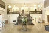 Located at 130 Brooke Ave in Norfolk, Virginia, this distinguished historic building exudes timeless architectural elegance throughout its single floor. The centerpiece of the space is a breathtaking grand lobby, where soaring ceilings draw the eye upward past elaborately detailed Corinthian columns adorned with ornamental capitals. Polished terrazzo floors reflect the warm glow of pendant globe lanterns suspended from above, lending the space a refined, luminous ambiance.

A sweeping curved staircase commands immediate attention, featuring striking black-and-white marble treads and an intricately crafted wrought iron balustrade with scrolling floral motifs. The upper mezzanine level, accessible via this dramatic staircase, overlooks the expansive main hall below. Decorative frieze work and classical moldings line the upper walls, reinforcing the building's historic character.

A glass-enclosed interior suite opens off the main lobby, revealing a modern kitchen and lounge area complete with a fireplace, chandelier, and contemporary furnishings — a refined contrast to the classical surroundings. Dual ADA-accessible drinking fountains are thoughtfully integrated into one of the corridors. Throughout, the interplay of historic architectural detail and updated interior elements creates a space that is both functionally versatile and architecturally significant.