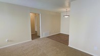 This single-story space presents an open layout that seamlessly connects the living and dining areas. The living area is finished with neutral-toned carpet, providing a comfortable and warm underfoot feel, while the dining area transitions to smooth hardwood-style flooring, creating a clear visual distinction between the two spaces. Neutral beige walls throughout contribute to a clean, understated aesthetic that works well as a blank canvas for personalization. A classic bowl-style pendant light hangs above the dining area, serving as a modest focal point and providing direct overhead illumination. The textured ceiling adds subtle visual interest. A floor vent is visible along the wall, indicating a forced-air HVAC system for climate control. An open doorway leads to a hallway that connects to additional rooms beyond, hinting at a functional and well-organized floor plan. The overall ambiance is straightforward and practical, with consistent neutral tones and modest finishes throughout. The space is move-in ready and offers a versatile foundation suitable for a variety of furnishing styles and arrangements.