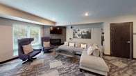This well-appointed community lounge is a single-floor space designed to accommodate a variety of social activities. The open layout integrates several distinct zones that flow seamlessly into one another, creating a dynamic and inviting atmosphere throughout.

At the heart of the space is a sleek bar area anchored by a striking blue mosaic tile backsplash. A white quartz countertop peninsula with rust-colored bar stools provides casual seating, while pendant lights in polished metallic finishes hang overhead. Behind the bar, a full-size refrigerator, sink, and dedicated coffee station ensure convenient refreshments.

Adjacent to the bar, a billiards table takes center stage on patterned carpet, flanked by a comfortable lounge area featuring a large sectional sofa, accent armchairs, and a stone-clad fireplace topped with a wall-mounted television — ideal for relaxed entertainment.

The dining nook offers a warm, intimate setting with a wraparound striped banquette, accent pillows, a solid wood table, and upholstered chairs beneath a trio of geometric pendant lights. Original artwork adorns the walls throughout.

A bright, windowed seating area with natural views of surrounding greenery provides additional dining tables and high-backed lounge chairs, making excellent use of natural light. Patterned carpet and hardwood flooring accents tie the entire space together with a cohesive, modern aesthetic.