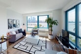This well-appointed waterfront residence is defined by its striking teal-framed floor-to-ceiling windows, which frame sweeping views of the river and city skyline from multiple rooms. Light wood flooring runs throughout, contributing to the bright, cohesive aesthetic that characterizes the entire space.

The living area is open and inviting, bathed in natural light from oversized windows that face the waterfront. The room comfortably accommodates a seating arrangement centered around a geometric area rug, with space for a bar cart and media console. Corner window placement in the living room provides panoramic sightlines across the river and skyline.

The kitchen is outfitted with high-gloss grey cabinetry, white quartz countertops, a subway-tile backsplash, and a full suite of stainless steel appliances including a range with hood ventilation and a dishwasher. Light wood flooring continues seamlessly from the main living area.

Both bedrooms feature the same signature teal-framed windows with direct waterfront and skyline views, filling each room with natural light. The primary bedroom includes a dedicated dresser area and wall-mounted lighting.

The primary bathroom offers a glass-enclosed walk-in shower with textured tile surround, a wide white quartz vanity, grey cabinetry, and a backlit mirror. A separate, clean-lined powder room serves as a secondary bathroom.